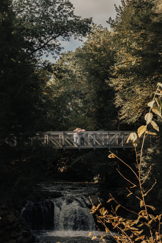 couple standing on bridge with waterfall underneath during their outdoor engagement photos at sweet arrow lake park in pine grove pennsyvania