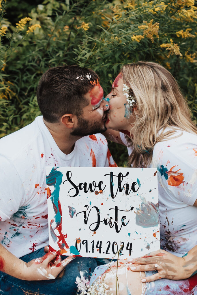couple kissing while holding save the date sign they painted during their outdoor engagement photo session