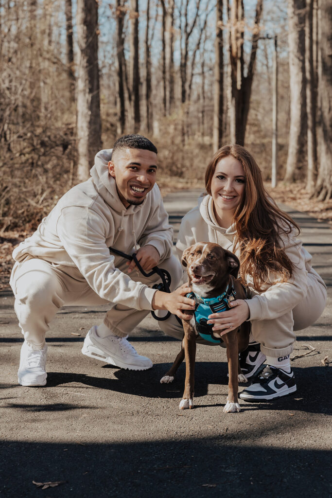 A couple walking hand-in-hand with their dog through a leafy park during their engagement session, capturing relaxed, natural moments.