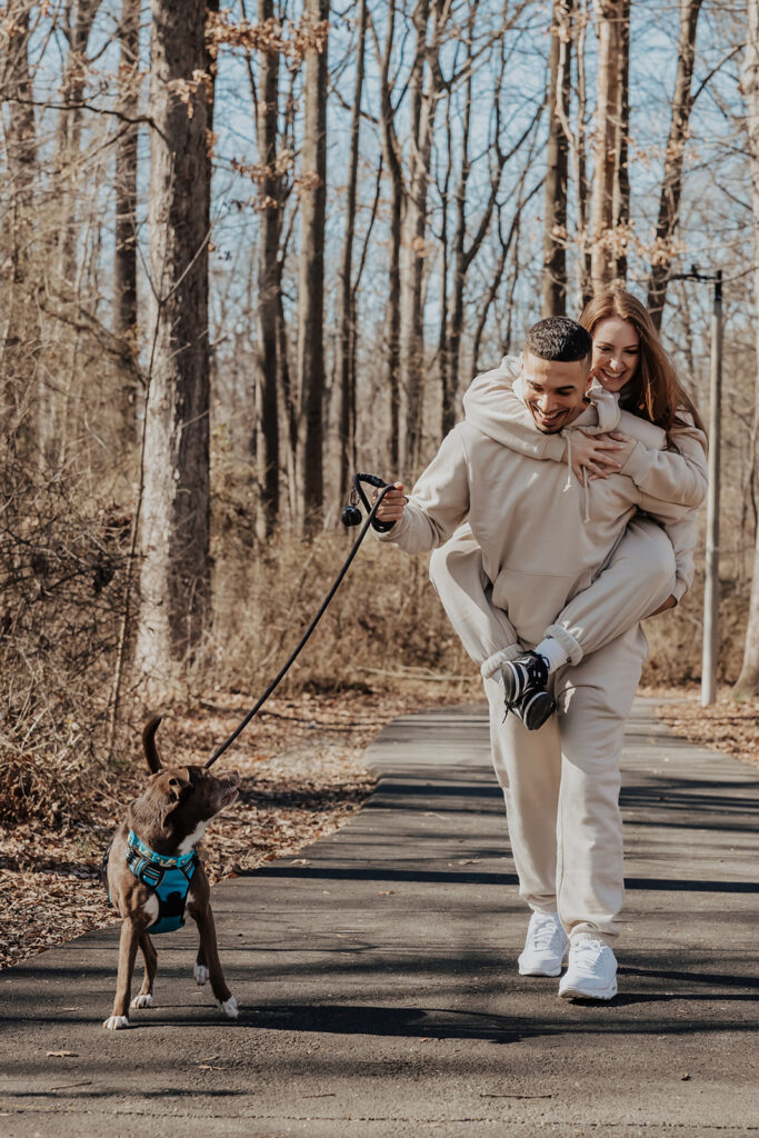 A couple walking hand-in-hand with their dog through a leafy park during their engagement session, capturing relaxed, natural moments.